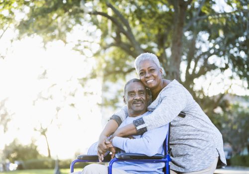 Portrait of a senior African American couple outdoors, showing their affection in the bright sunlight.  The man is sitting in a wheelchair, in the warm embrace of his devoted wife.  They are smiling at the camera.