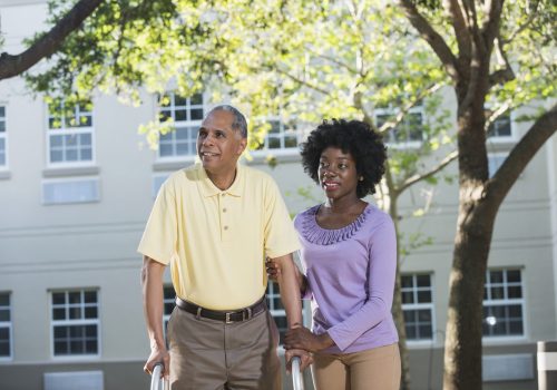 A young African American woman helping her father using a walker. They are outdoors, a building in the background. The man is in his 50s. His daughter is standing beside him, holding his arm. They are smiling looking away from the camera.