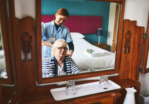 Reflection of happy young woman healthcare worker combing hair of a senior female patient at retirement home